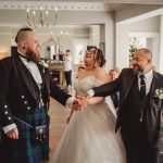 Bride and two men holding hands, smiling at wedding.