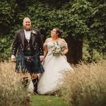 Couple in wedding attire walking through a meadow.