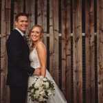 Smiling couple in wedding attire by wooden fence.