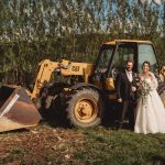 Bride and groom standing by yellow construction vehicle.