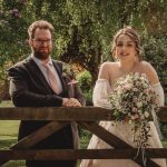 Bride and groom smiling by garden fence, wedding day.