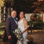 Smiling couple poses in garden by fountain, wedding attire.