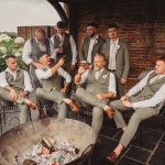 Groomsmen in grey suits sitting around outdoor fire pit.