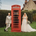 Bride and groom by red phone box, countryside setting.