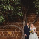 Couple poses by brick wall with tree backdrop.