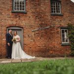 Couple in wedding attire outside brick cottage.