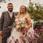 Bride and groom posing with floral bouquet in garden.