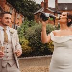 Bride drinking champagne, groom holding glasses.