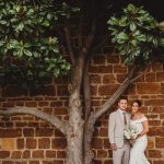 Bride and groom pose under tree against brick wall.