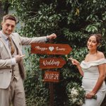 Couple posing by wooden wedding sign, greenery backdrop.