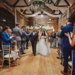 Bride and groom walk down wedding aisle, guests clapping.