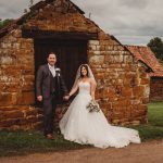 Bride and groom pose by rustic stone building.