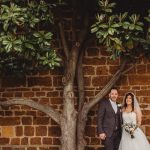 Bride and groom standing by brick wall and tree.