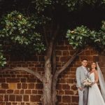 Wedding couple under tree against brick wall.