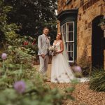 Wedding couple standing by stone building, lush garden.