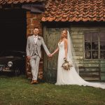 Couple in wedding attire outside rustic building.