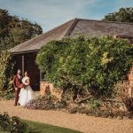 Bride and groom by brick building, sunny garden setting.