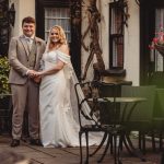 Bride and groom posing outdoors by chairs, smiling.