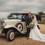 Bride and groom beside vintage car outdoors.