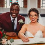 Smiling couple signing wedding registry with flowers nearby.