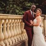 Smiling couple in wedding attire by stone balustrade.