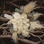 White roses and pampas grass bouquet on wooden chair.