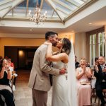 Newlyweds kiss at their wedding ceremony, guests applauding.