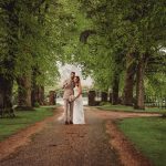 Wedding couple walking under trees with umbrella.