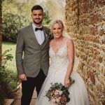 Smiling couple in wedding attire by stone wall.