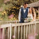 Couple smiling on wooden bridge in garden setting.