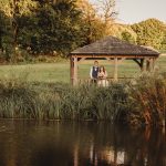 Couple in gazebo by pond, lush greenery surrounds.