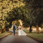 Bride and groom walking under tree canopy on path.