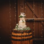 Rustic wedding cake on wooden barrel with lights.