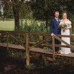 Bride and groom standing on wooden bridge outdoors.