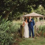 Couple in wedding attire by wooden gazebo outdoors.