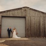 Couple holding hands, leaning on a rustic barn.