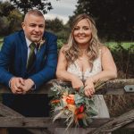 Wedding couple posing by wooden fence with flowers.